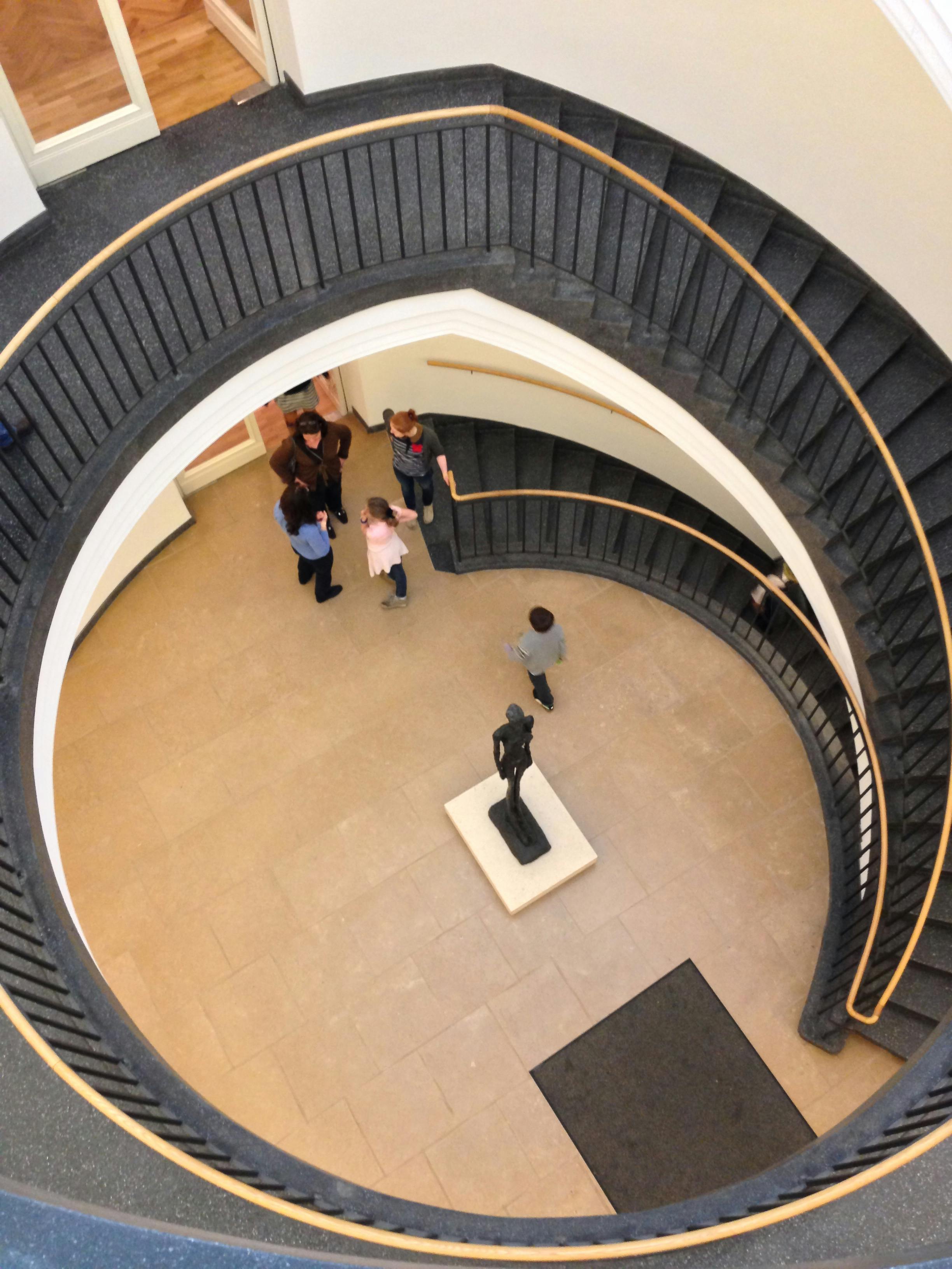 Άνθρωποι που στέκονται σε συνομιλία στη βάση μιας σπειροειδούς σκάλας με ένα άγαλμα στο κέντρο του ορόφου.