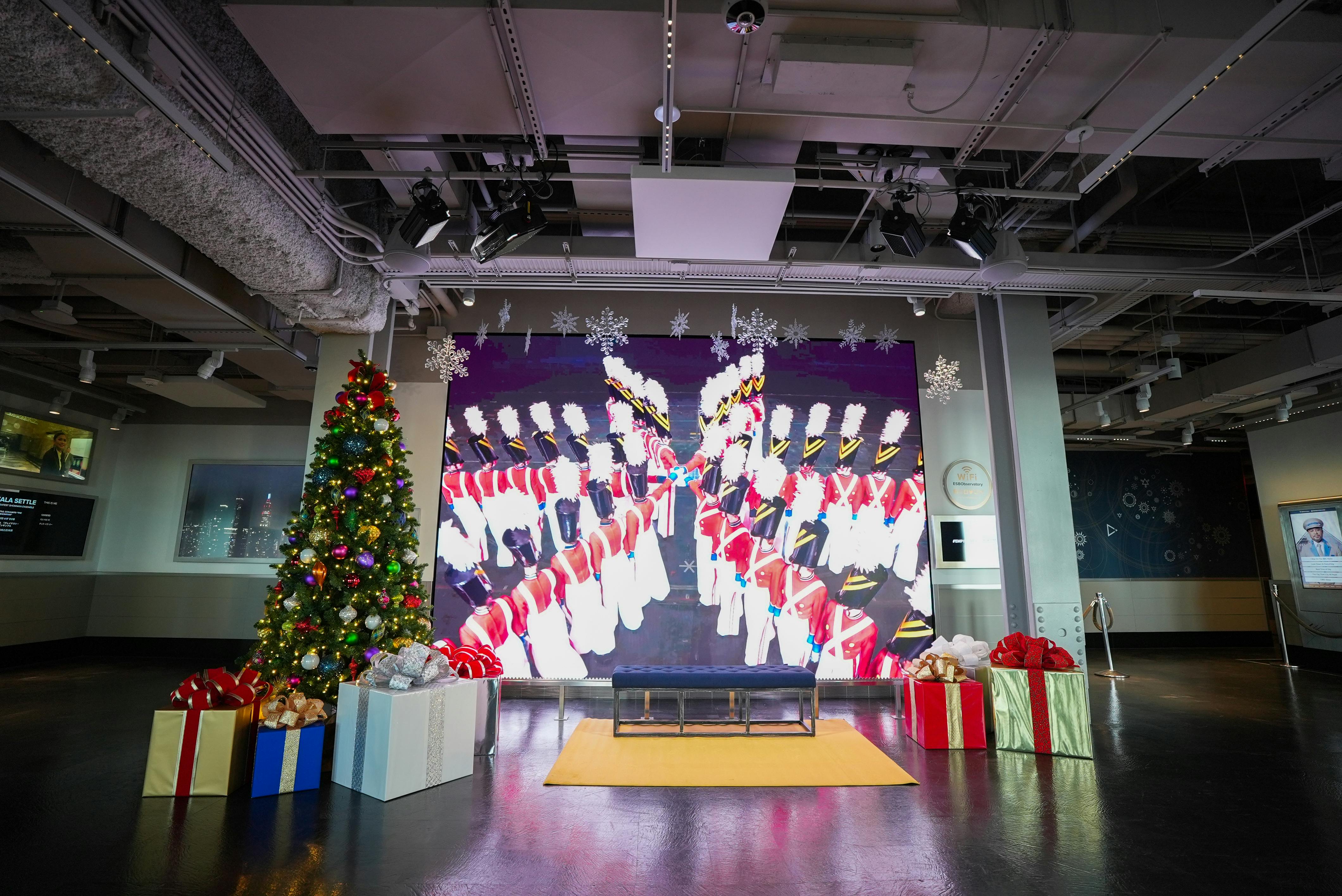 A festive indoor scene with a decorated Christmas tree, gift boxes, a seating bench, and a large screen showing colorful imagery.
