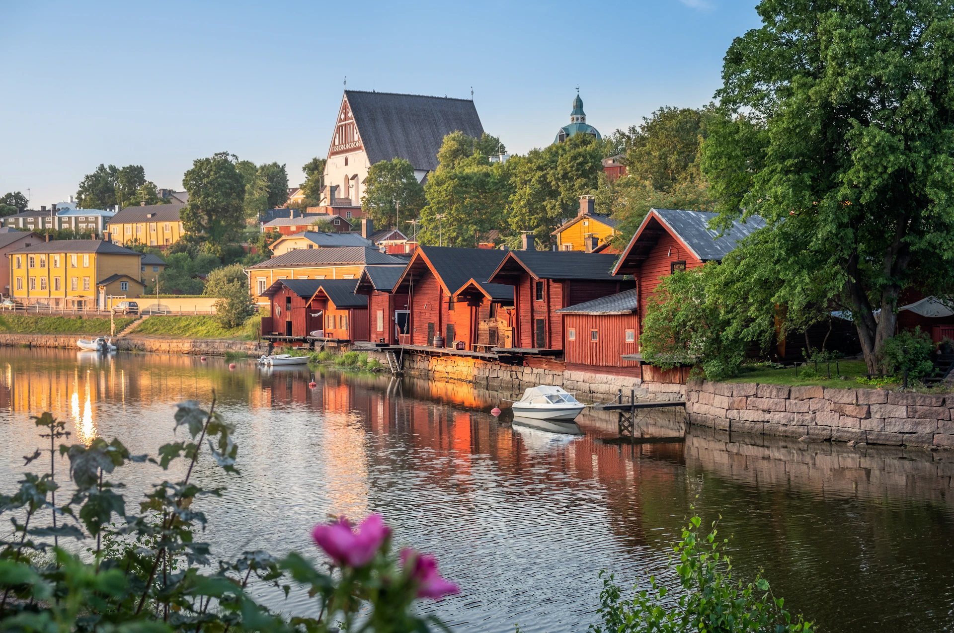 A scenic riverside view with red wooden buildings, moored boats, lush trees, and a large historic church on a hill in the background.