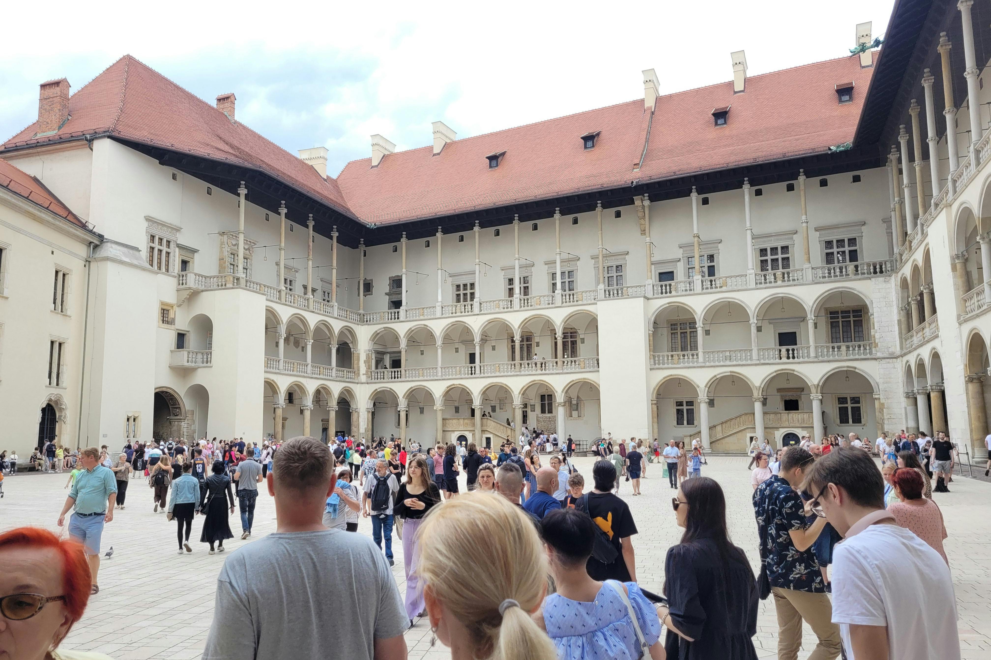 부분적으로 흐린 하늘 아래 아치가 있는 건물과 붉은 기와 지붕으로 둘러싸인 수많은 사람들이 있는 마당입니다.