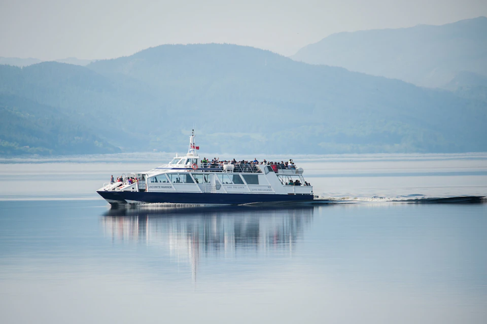 A ferry with passengers on board sails on a calm lake, with mountains visible in the background.