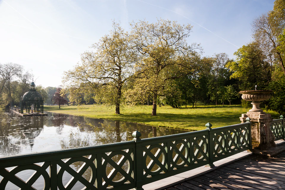 A peaceful park scene with tall trees, a pond, and a decorative green fence in the foreground under a clear blue sky.