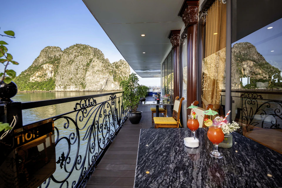 A scenic balcony overlooking water and cliffs, with a table holding two beverages and chairs by a glass wall.