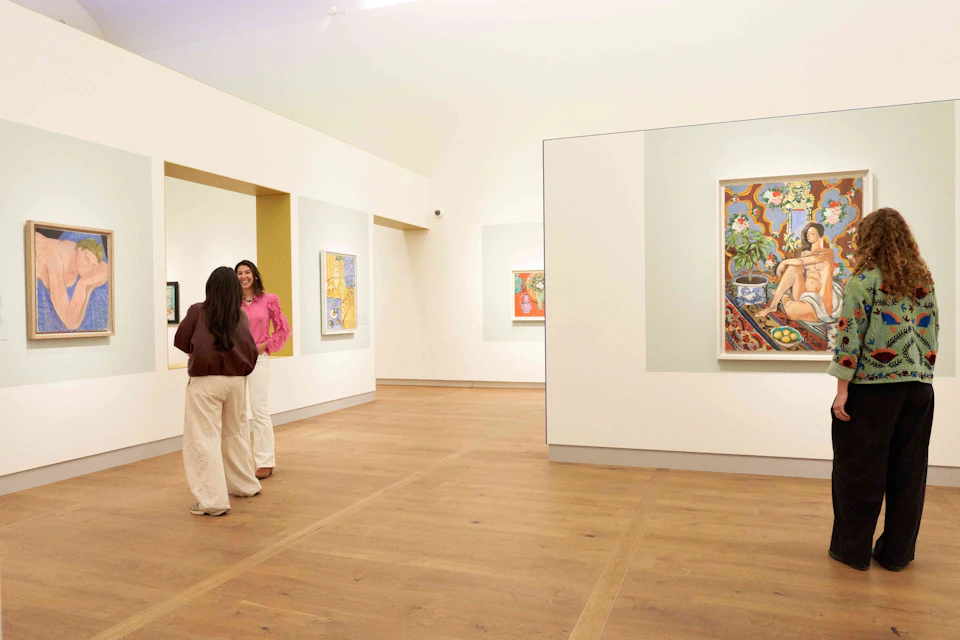 Two people stand in an art gallery with wooden floors, viewing colorful paintings displayed on the white walls.