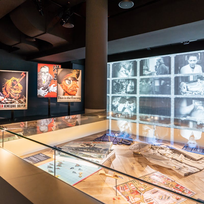 Un'esposizione museale con manifesti di propaganda della Seconda Guerra Mondiale, una vetrina con manufatti e una parete di immagini in bianco e nero proiettate.