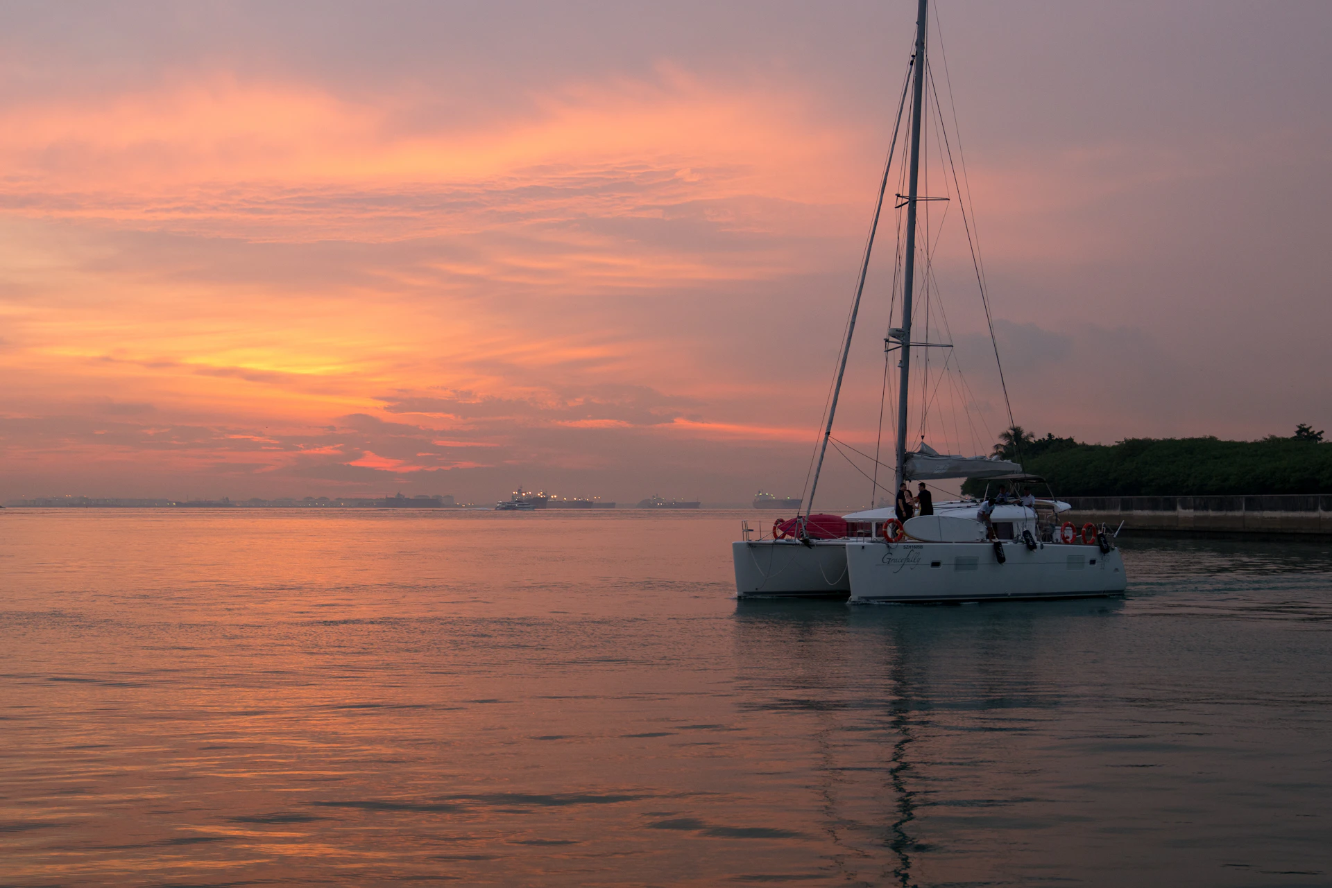 A sailboat on calm water during a pink-orange sunset, with distant ships on the horizon.