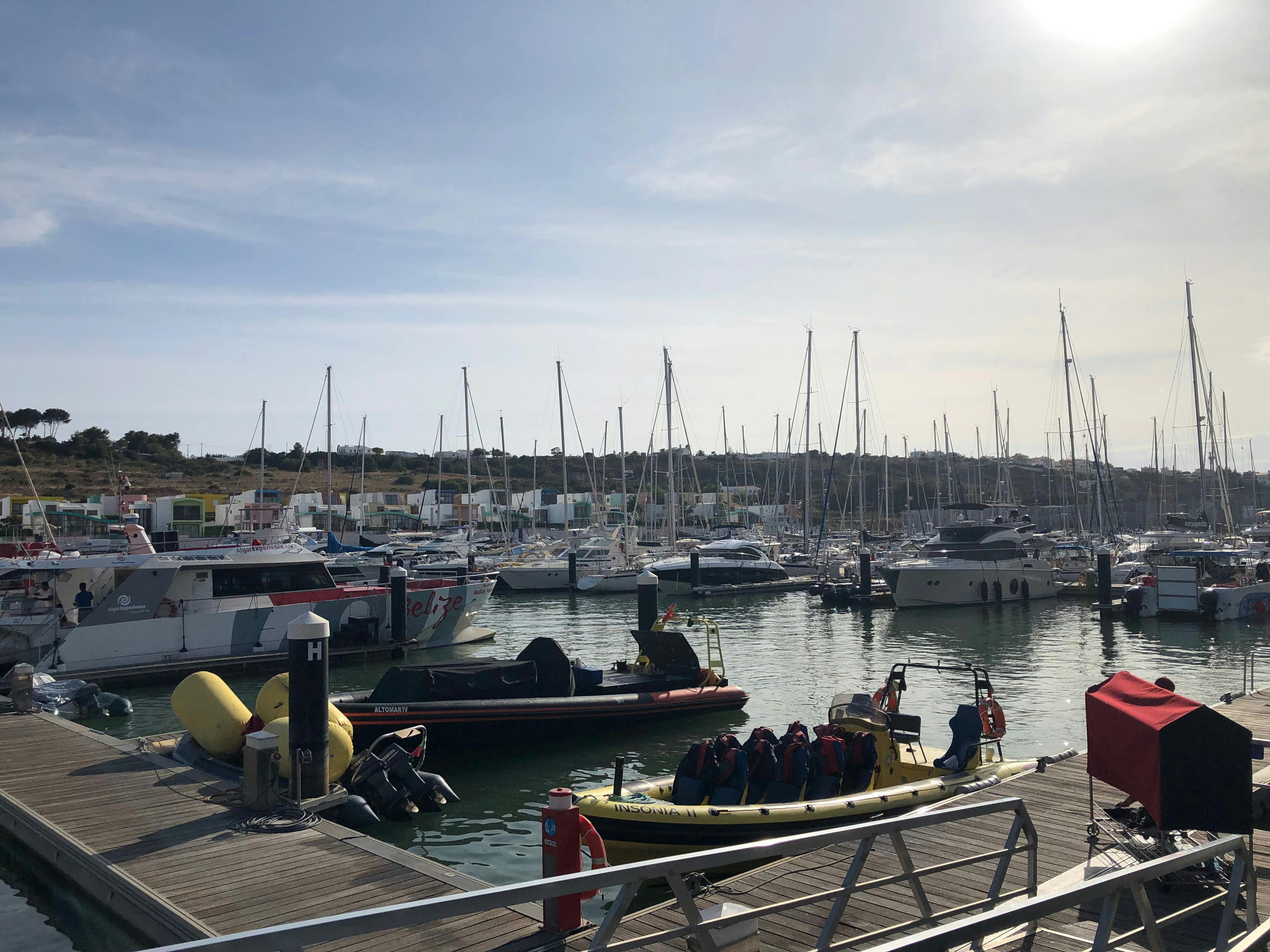 A marina with numerous boats and yachts docked, surrounded by hills and buildings, under a clear sky with the sun shining.