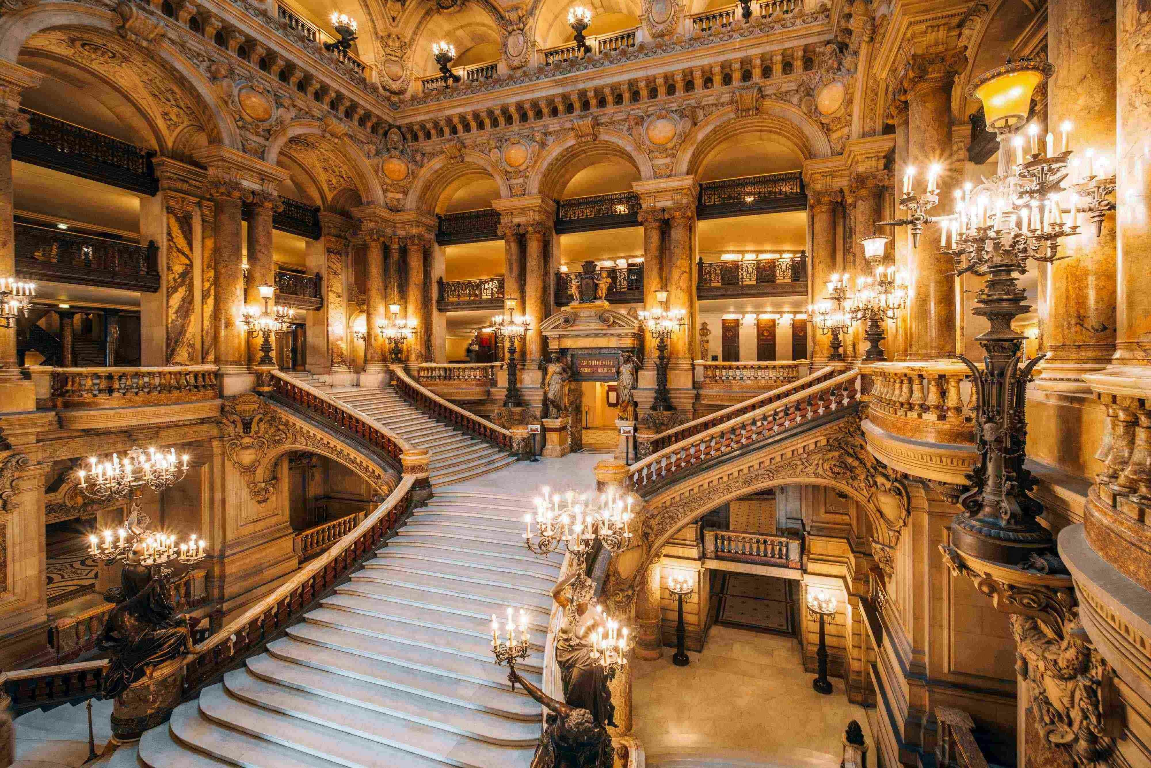 A grand staircase in an ornate hall with chandeliers, decorative columns, and intricate architectural details.