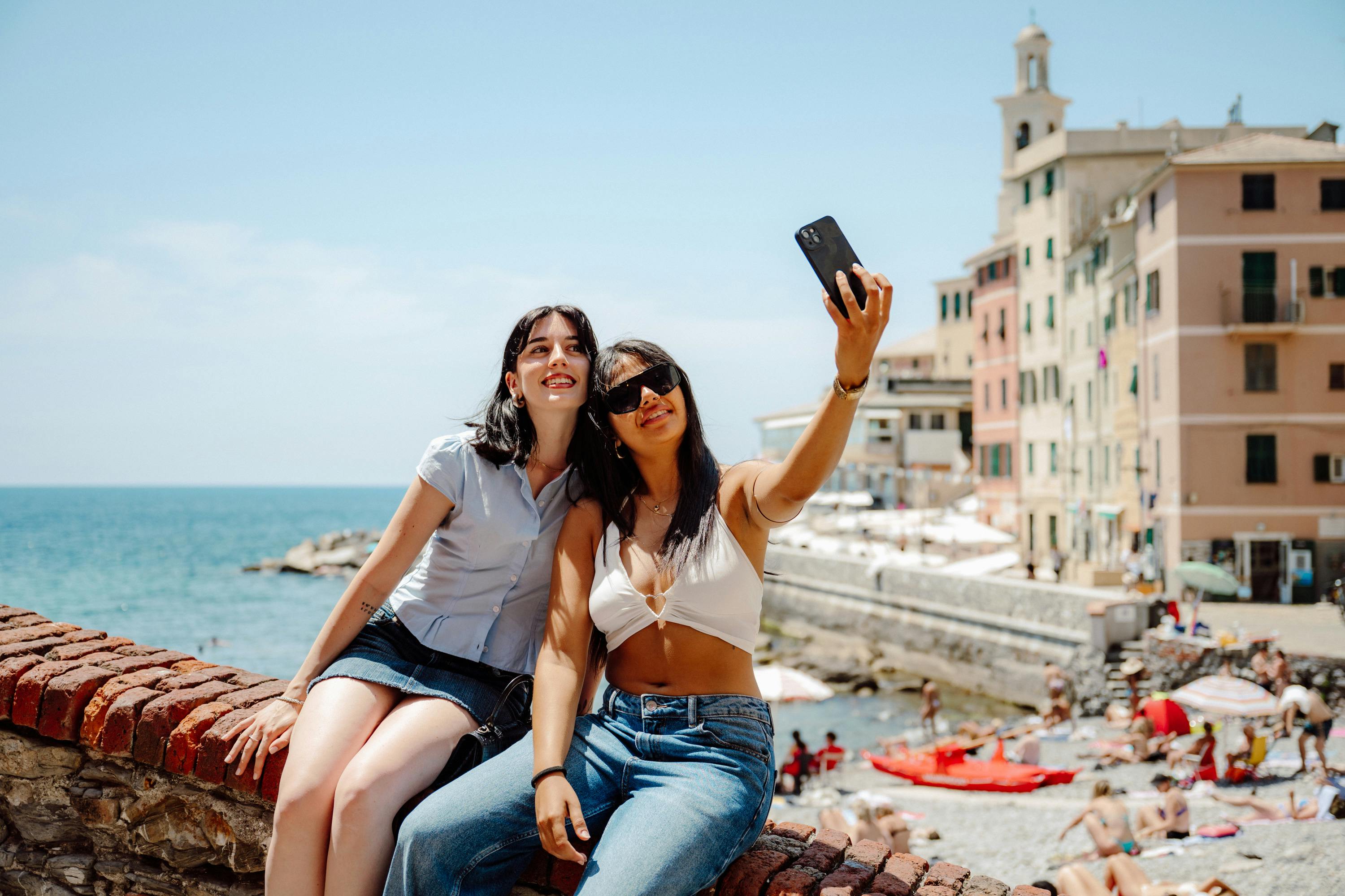 Dues dones fent-se una foto al costat d'una ciutat costanera, amb una paret de maons, mar i edificis al fons en un dia assolellat.