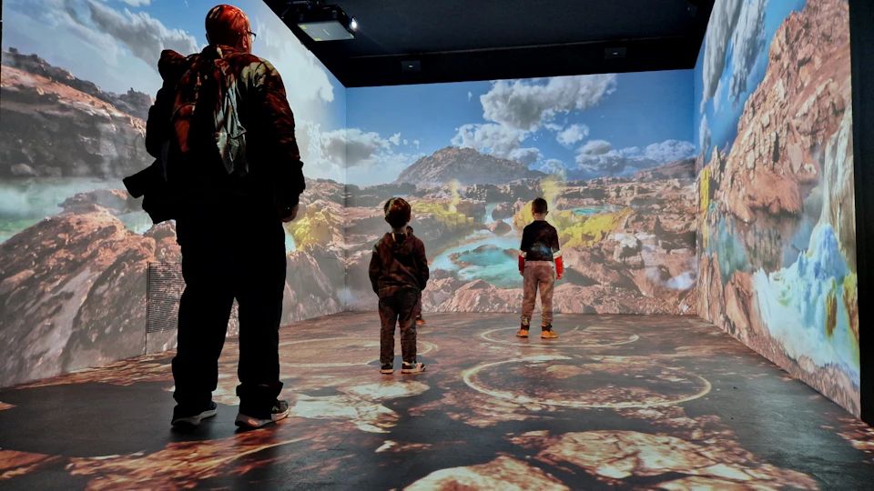 Three individuals, including two children, stand in a room with a detailed desert landscape projected on the walls and floor.