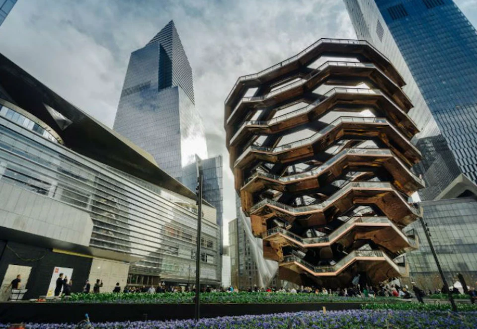 Modern urban plaza with unique honeycomb structure, tall skyscrapers, greenery, and people walking around. Sky is partly cloudy.