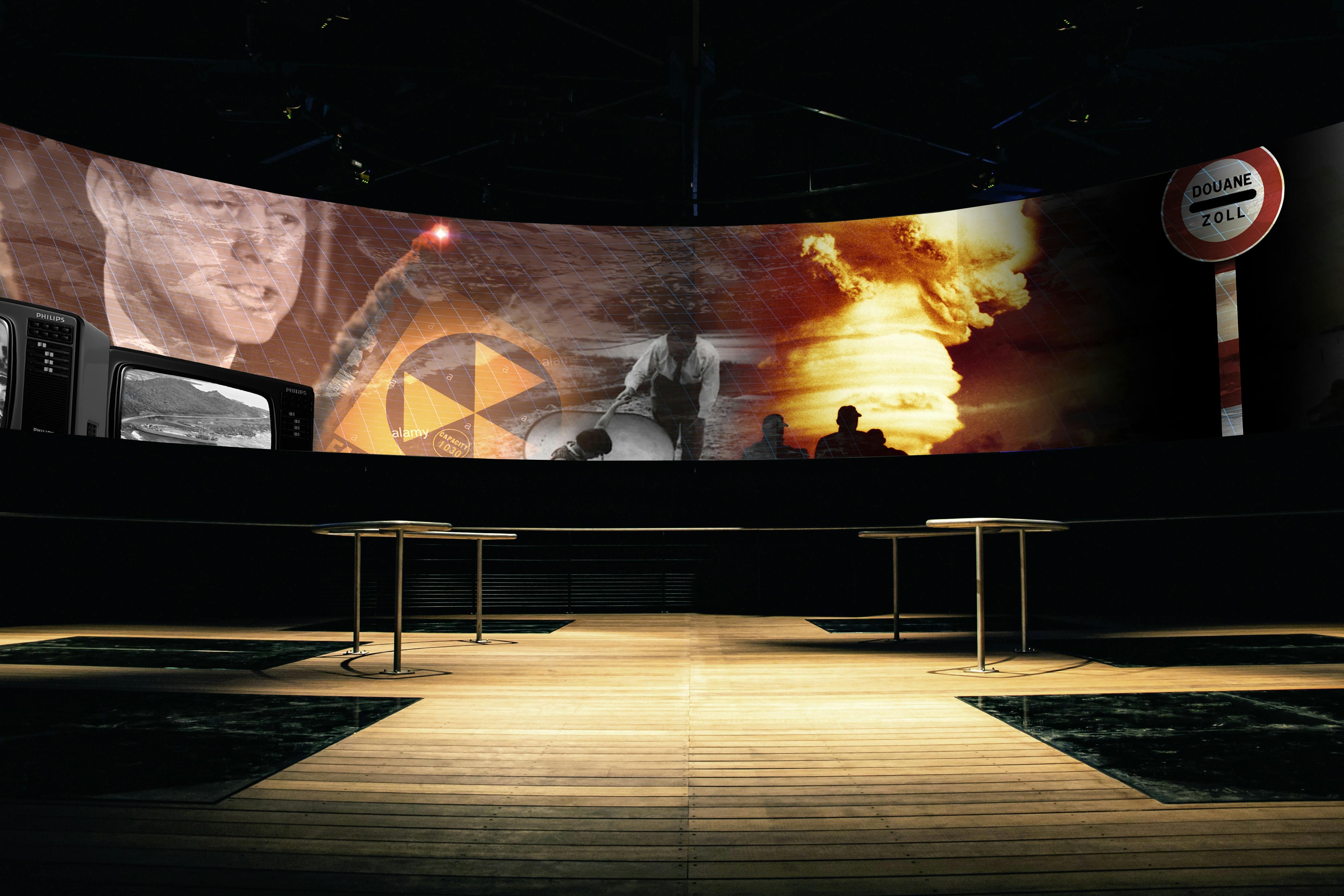 Schermo curvo che visualizza varie immagini, tra cui un uomo, un simbolo nucleare e un missile, in una stanza con pavimenti in legno e tavoli vuoti.
