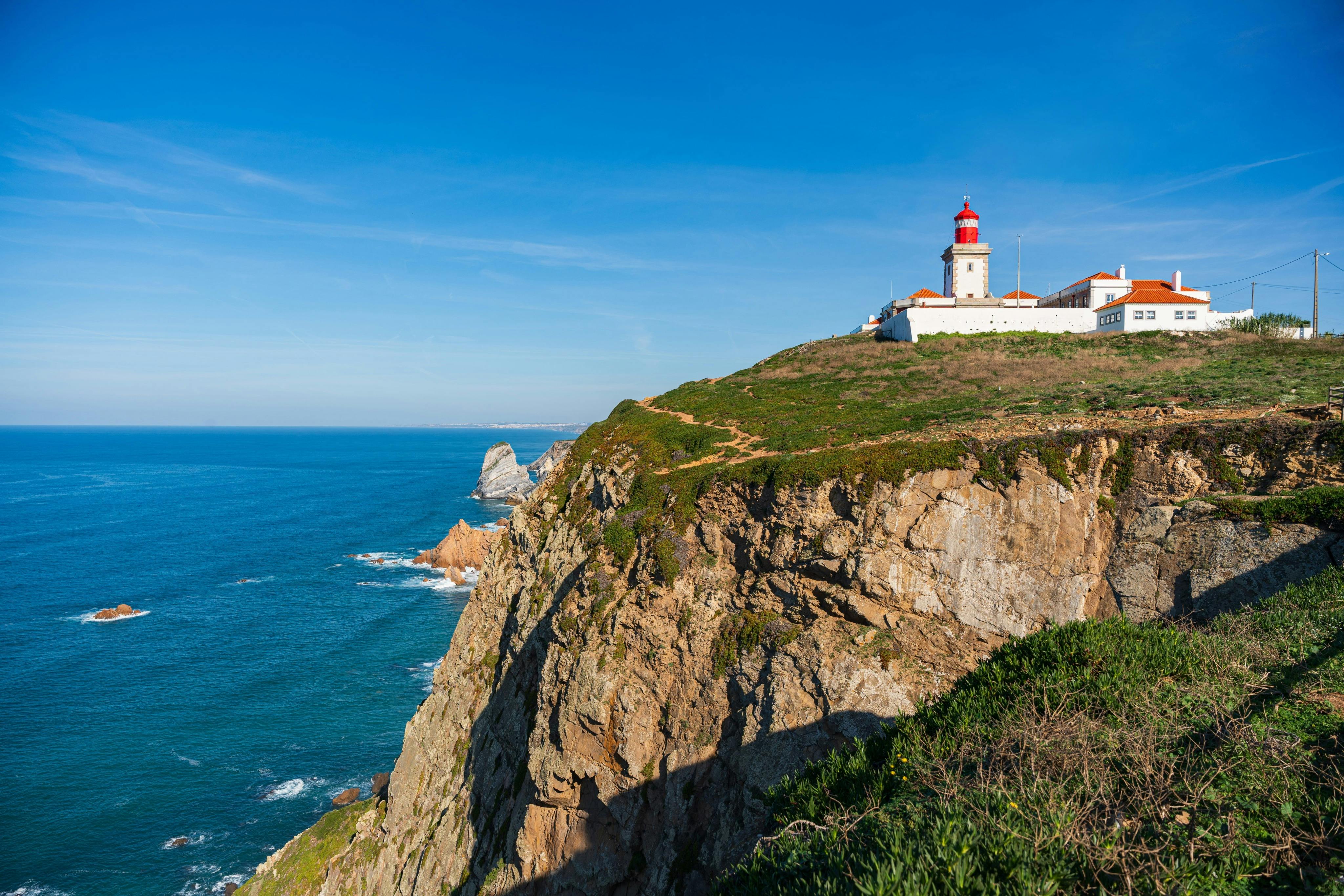 A white lighthouse with a red top on a green cliff overlooking the ocean under a clear blue sky.