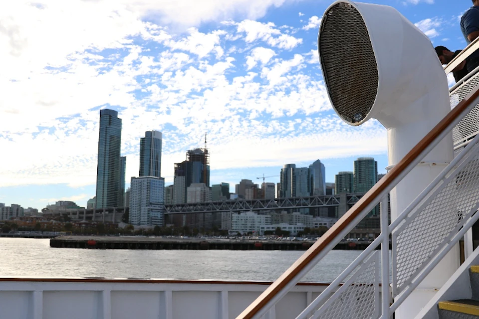 View of a city skyline with tall buildings and a bridge, as seen from a boat deck with railings and a large loudspeaker.