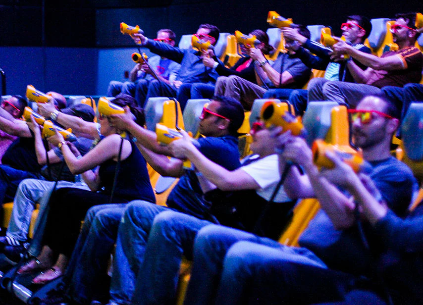 People seated in rows wearing 3D glasses, holding yellow guns, and aiming at screens in an interactive attraction.