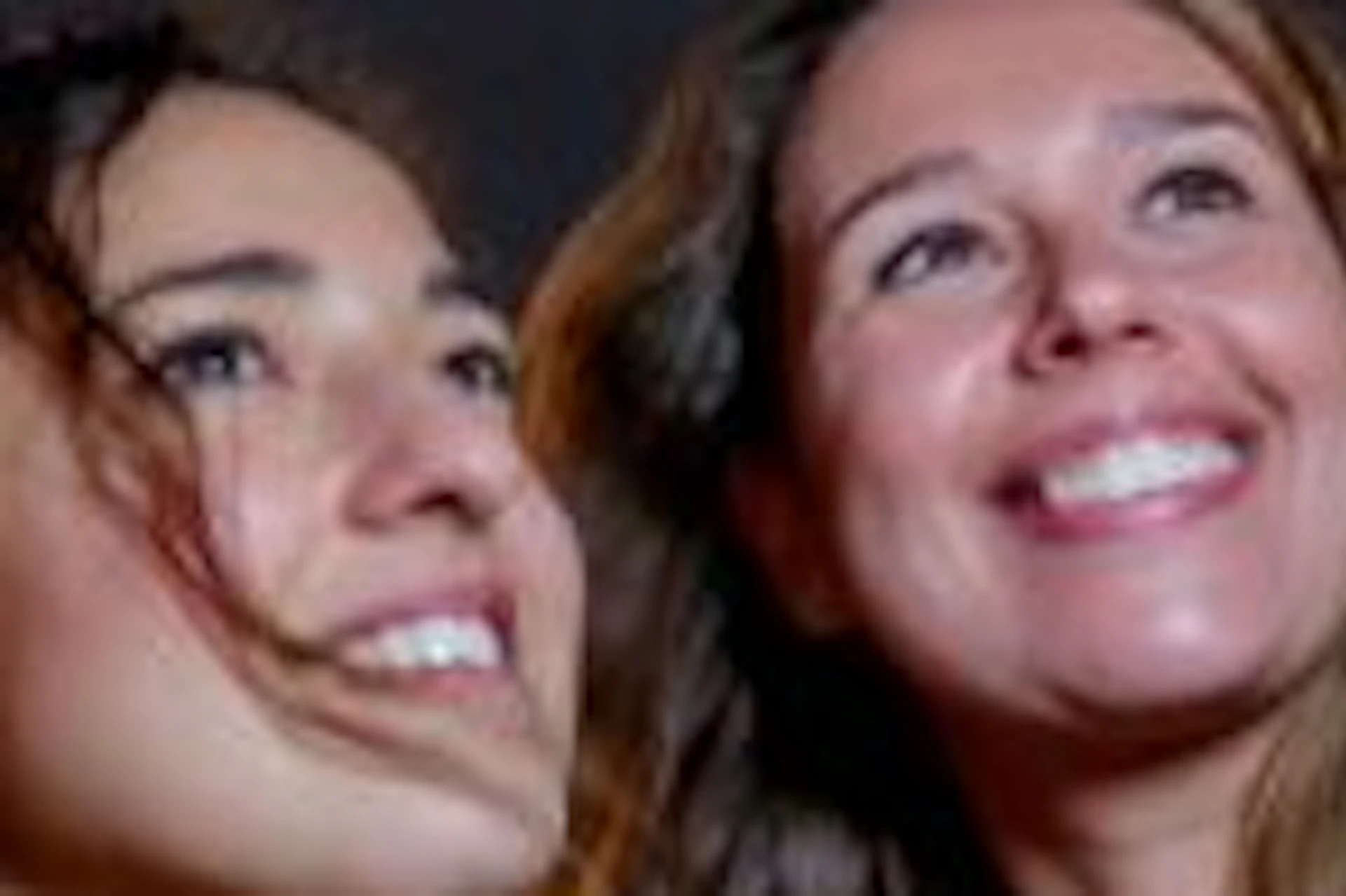 Two women take a selfie at night in front of lit buildings and a bridge over water.