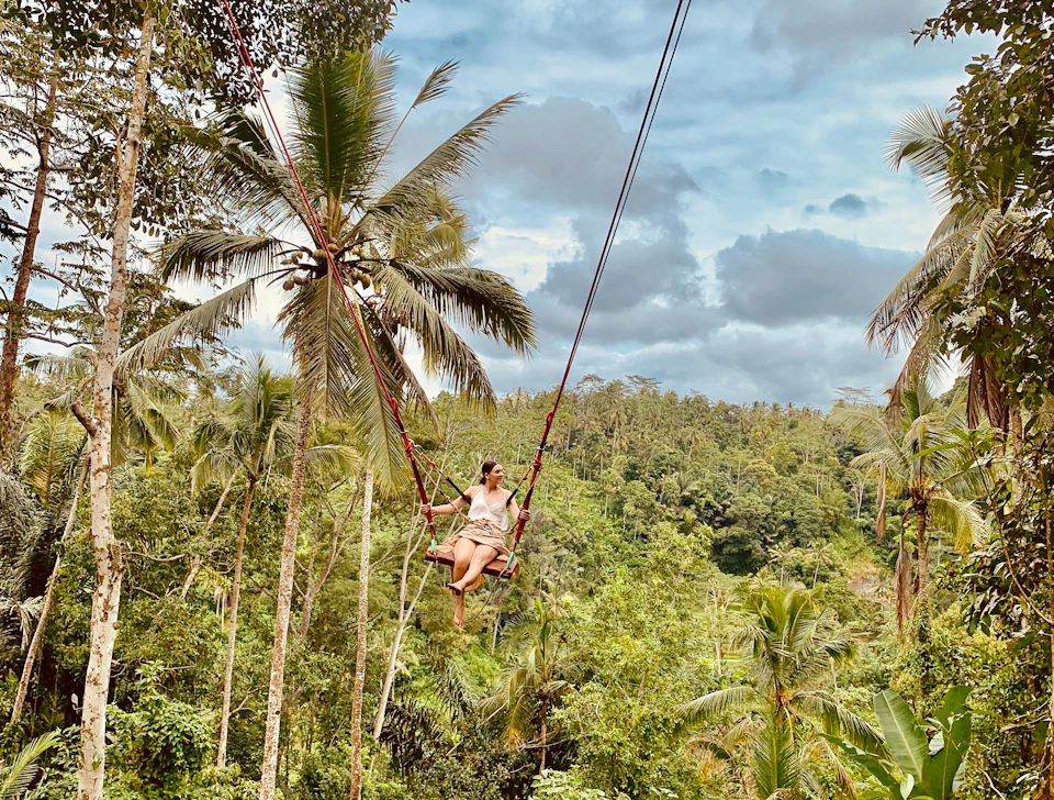 A person swings high above a lush tropical forest with palm trees under a partly cloudy sky.