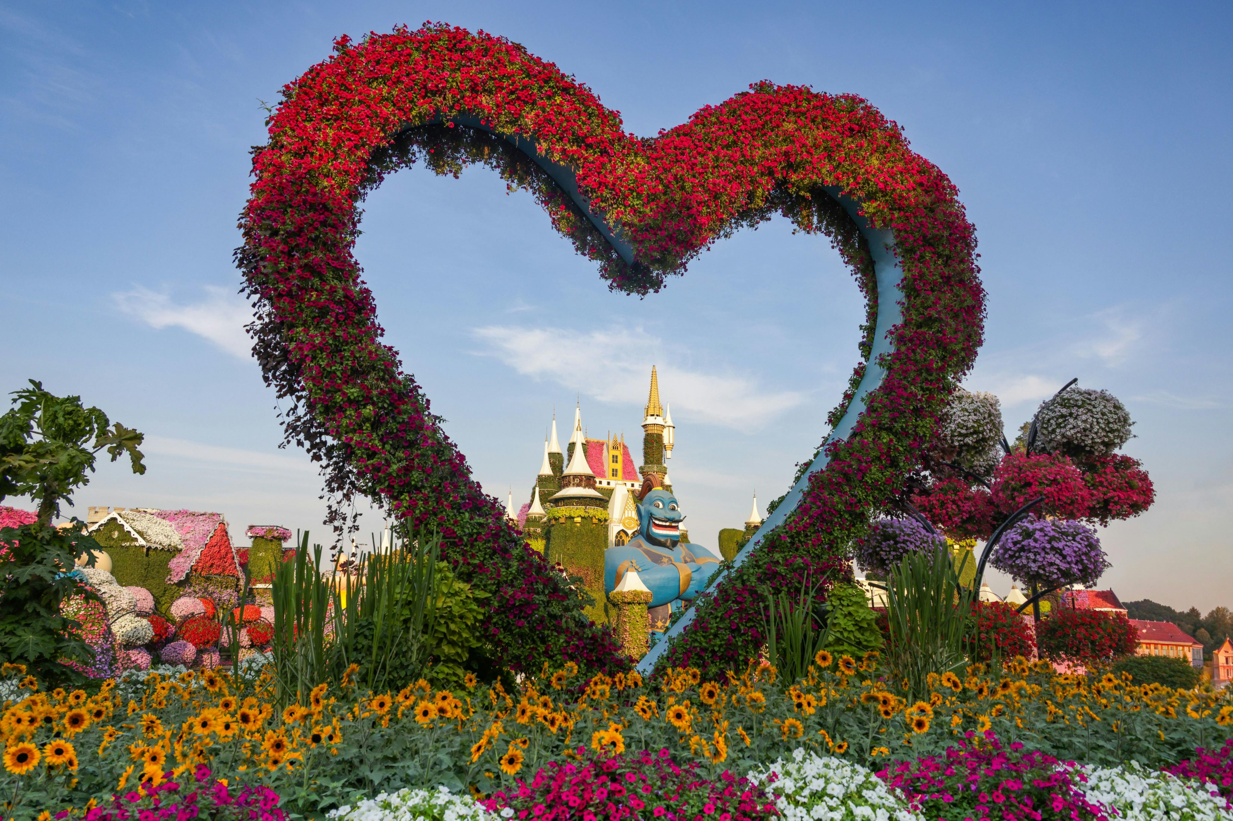 A large heart-shaped floral sculpture framing a castle in the background, surrounded by colorful flowers, under a clear sky.