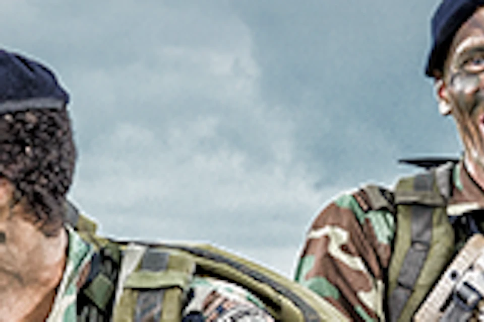 Six soldiers in camouflage uniforms and berets, holding rifles, crouching and smiling on a sandy beach under a cloudy sky.