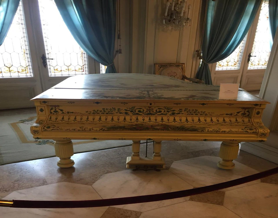 An ornate vintage piano in an elegant room with teal curtains, a chandelier, and a sign placed on the piano's lid.
