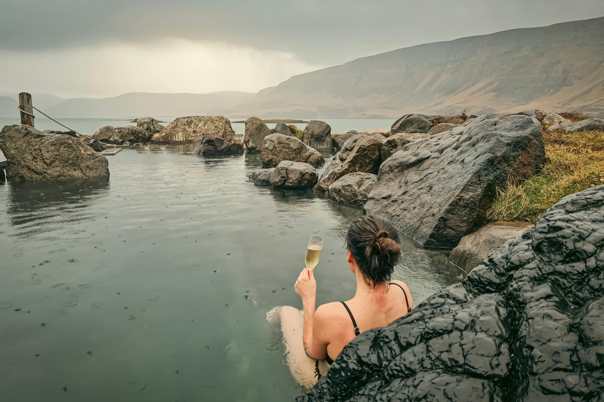 一位身穿黑色泳装的人在岩石温泉中举着一杯香槟酒，山上有雾气。