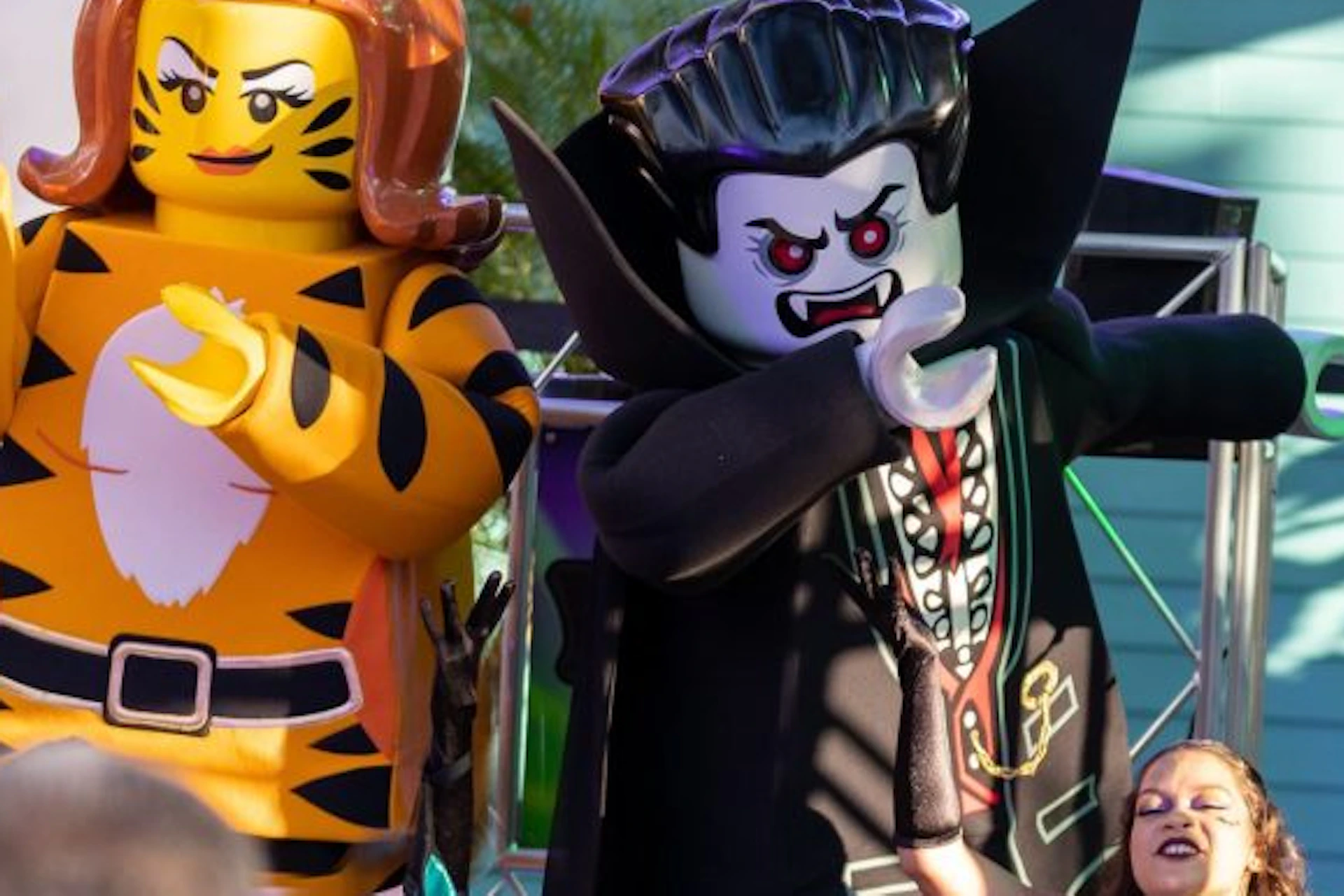 Large LEGO figures of a tiger and a vampire, surrounded by costumed people and stage lights, with smoke in the background.