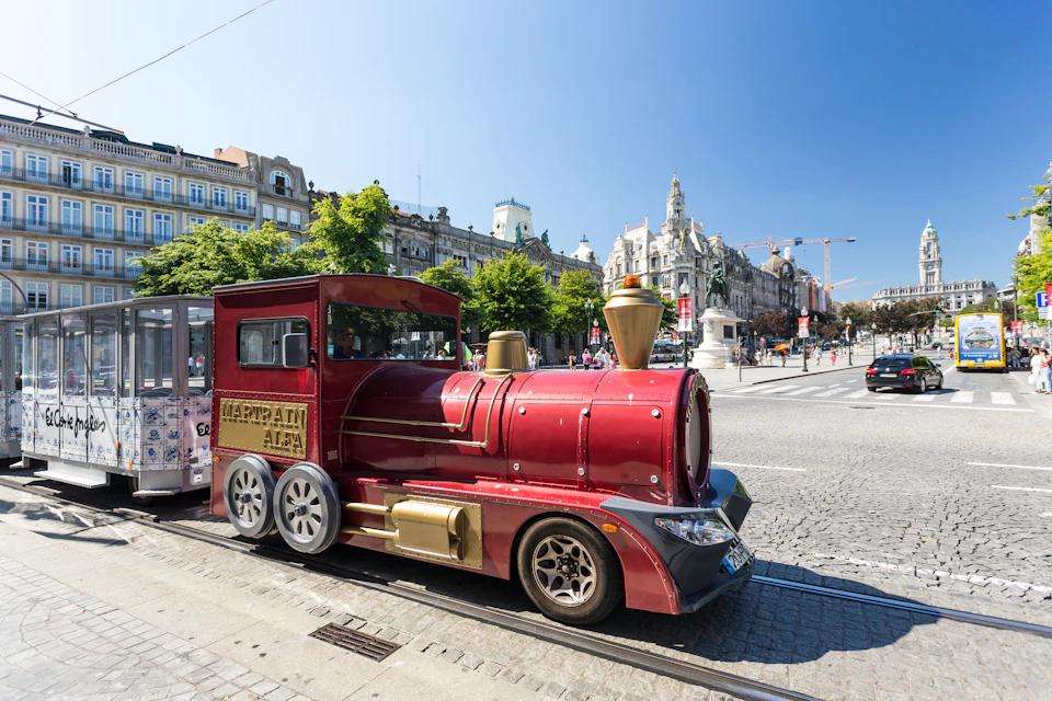 A small, red and gold tourist train parked on a cobblestone street in a European city with historic buildings in the background.