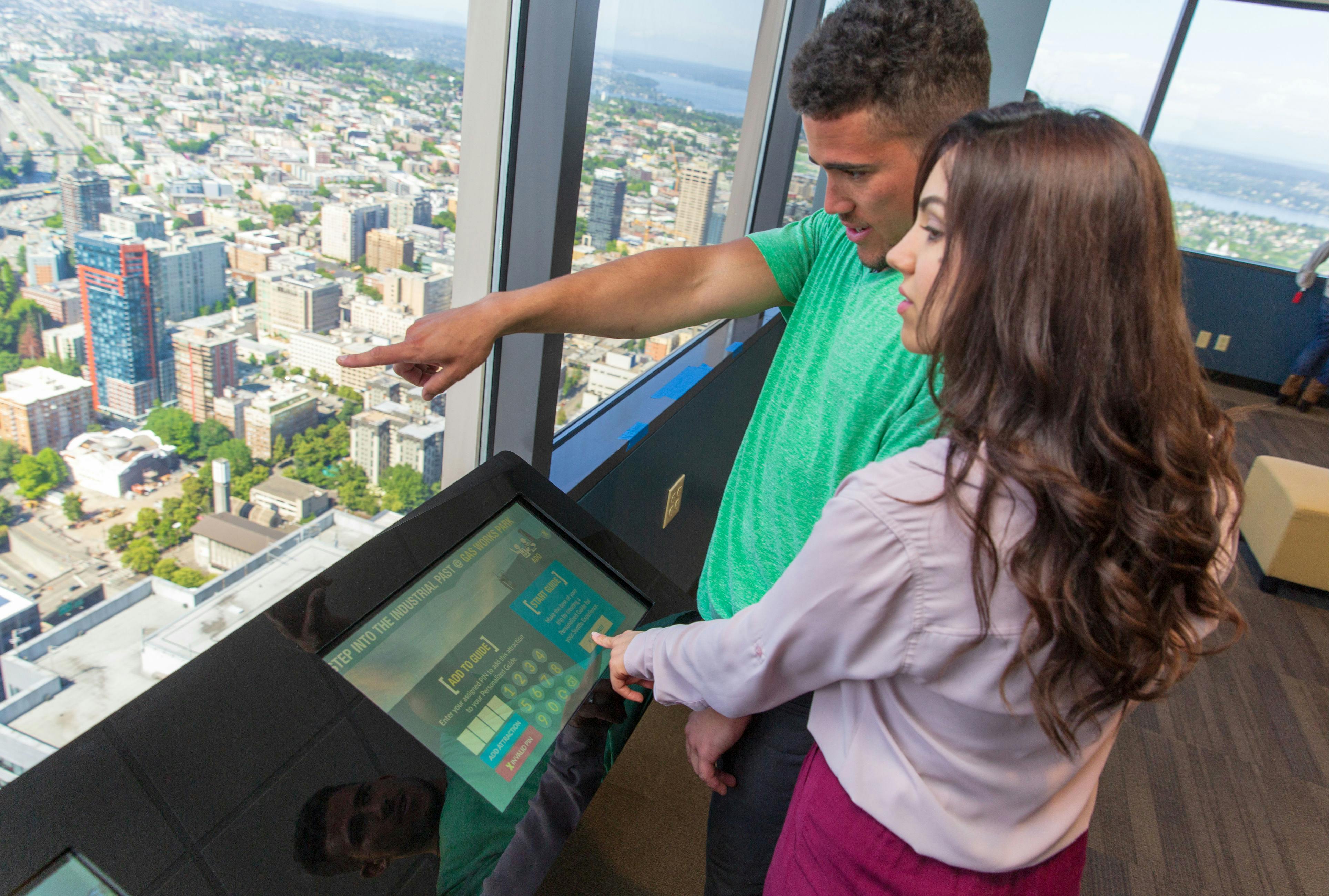 Два человека взаимодействуют с информационным киоском в высотном здании с большими окнами, откуда открывается вид на город.