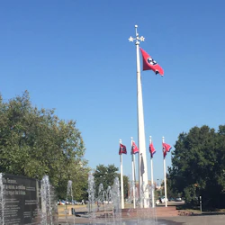 A plaza with a fountain, flagpoles displaying Tennessee flags, a memorial wall, and trees under a clear blue sky.
