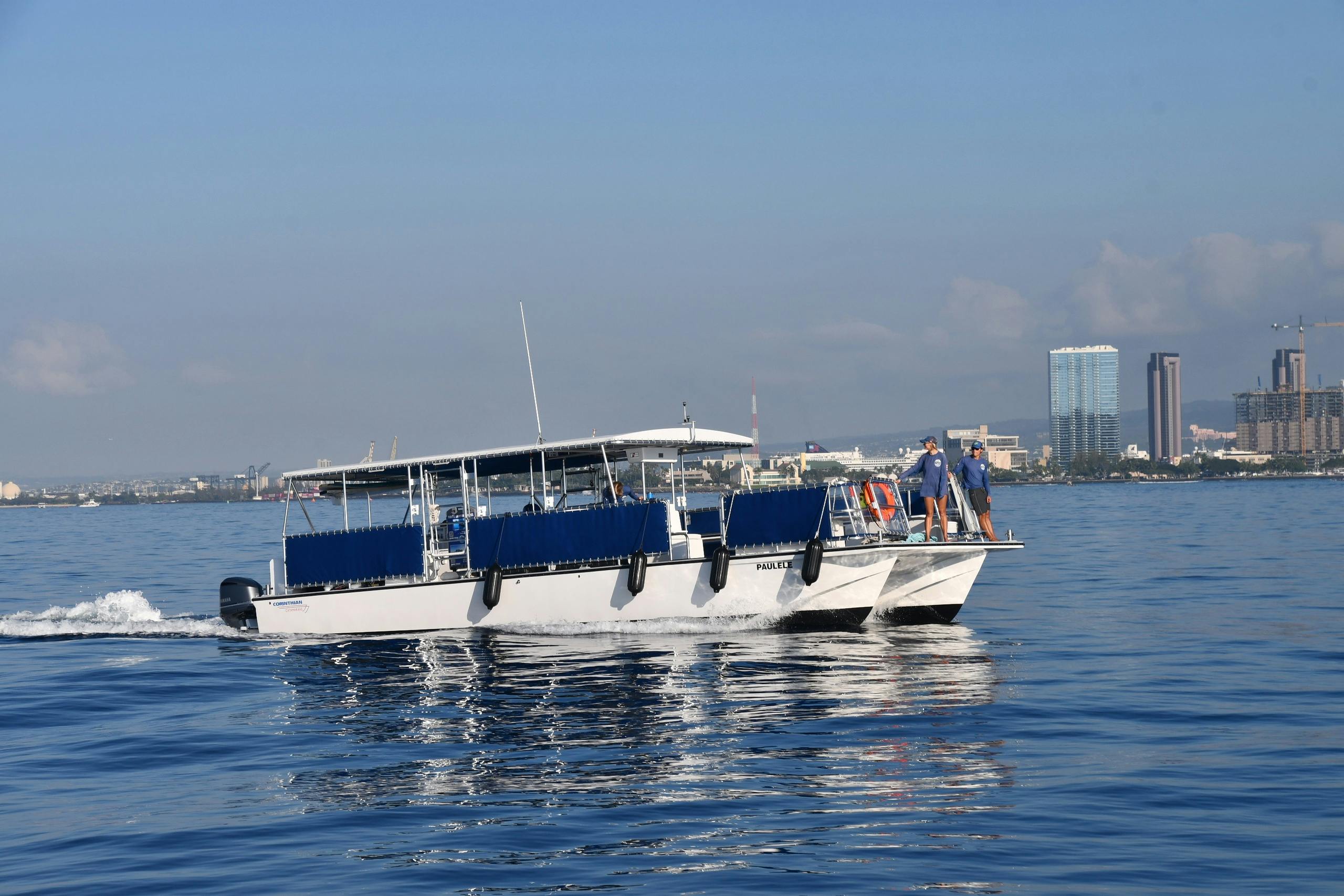 Un barco de pasajeros blanco con asientos azules está en aguas tranquilas, con gente a bordo y un paisaje urbano visible al fondo.