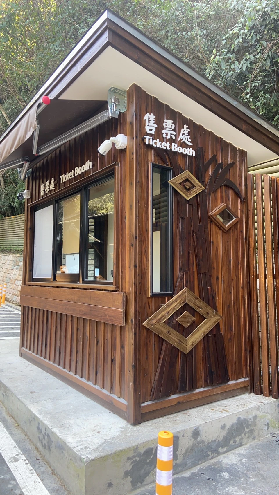 A wooden ticket booth with decorative elements and signs in English and Chinese, located outdoors near a path and trees.
