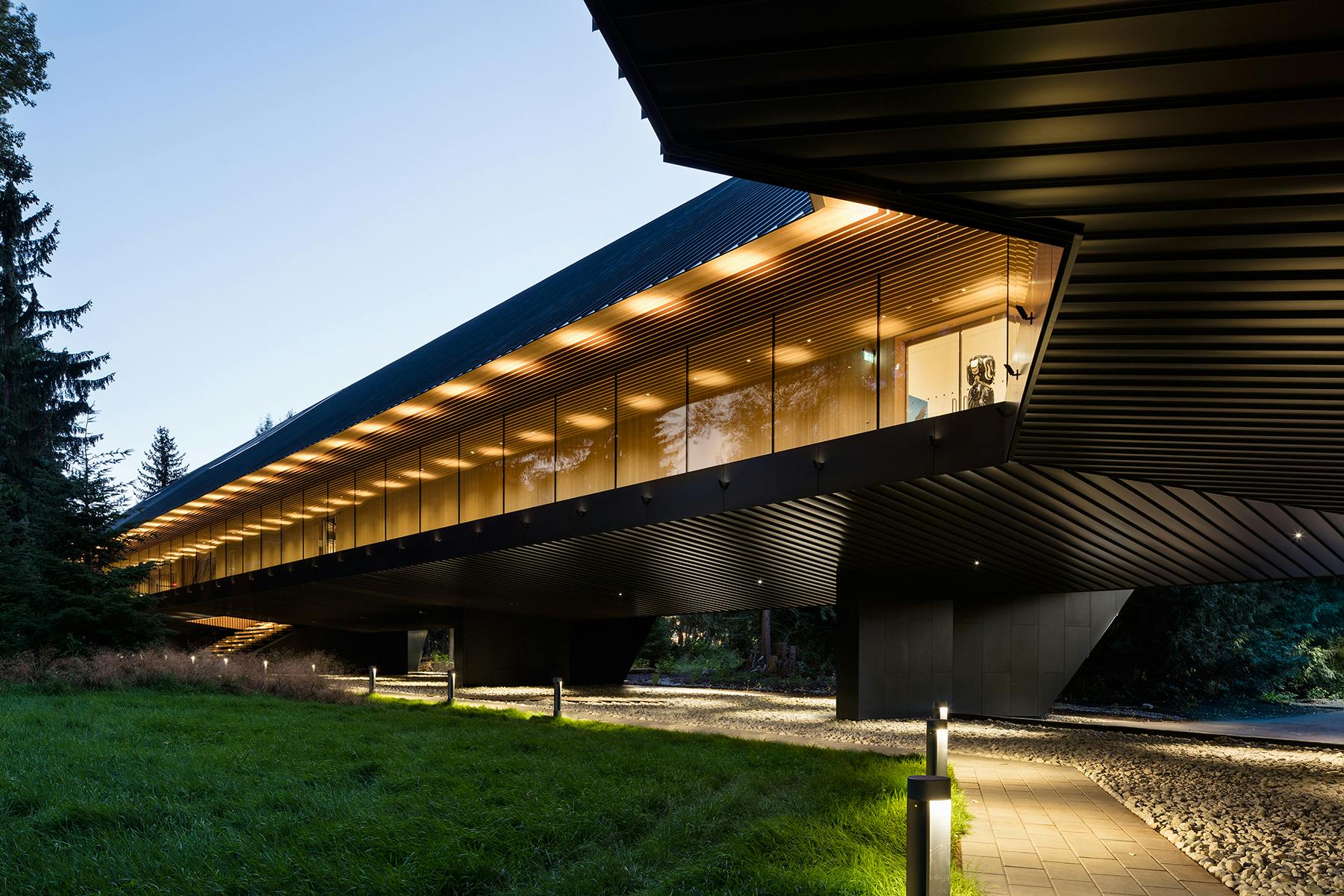 Un bâtiment moderne avec de grandes fenêtres en verre et des lumières extérieures, surélevé au-dessus d'une zone herbeuse avec un sentier.