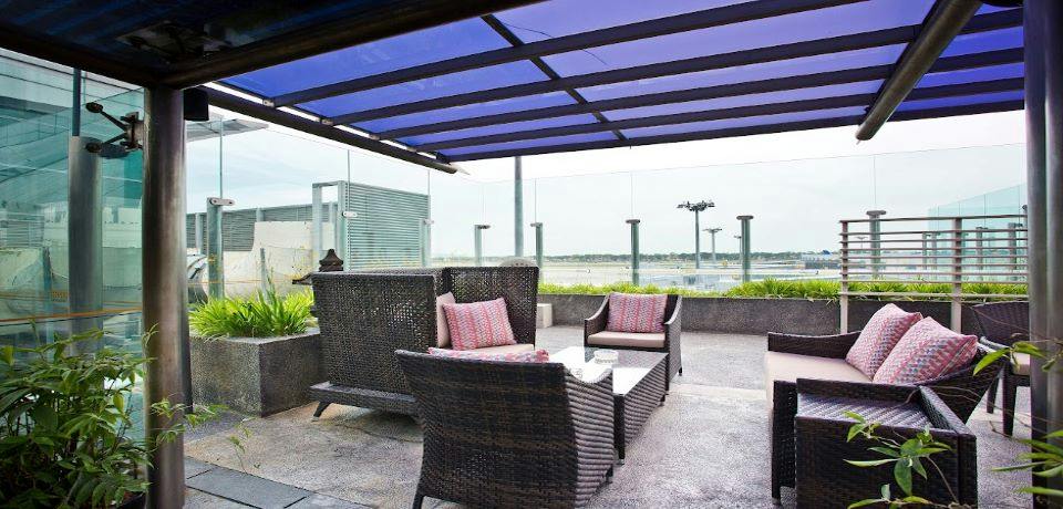 Outdoor patio area with wicker furniture, cushions, a glass table, and a blue pergola roof against a clear sky backdrop.