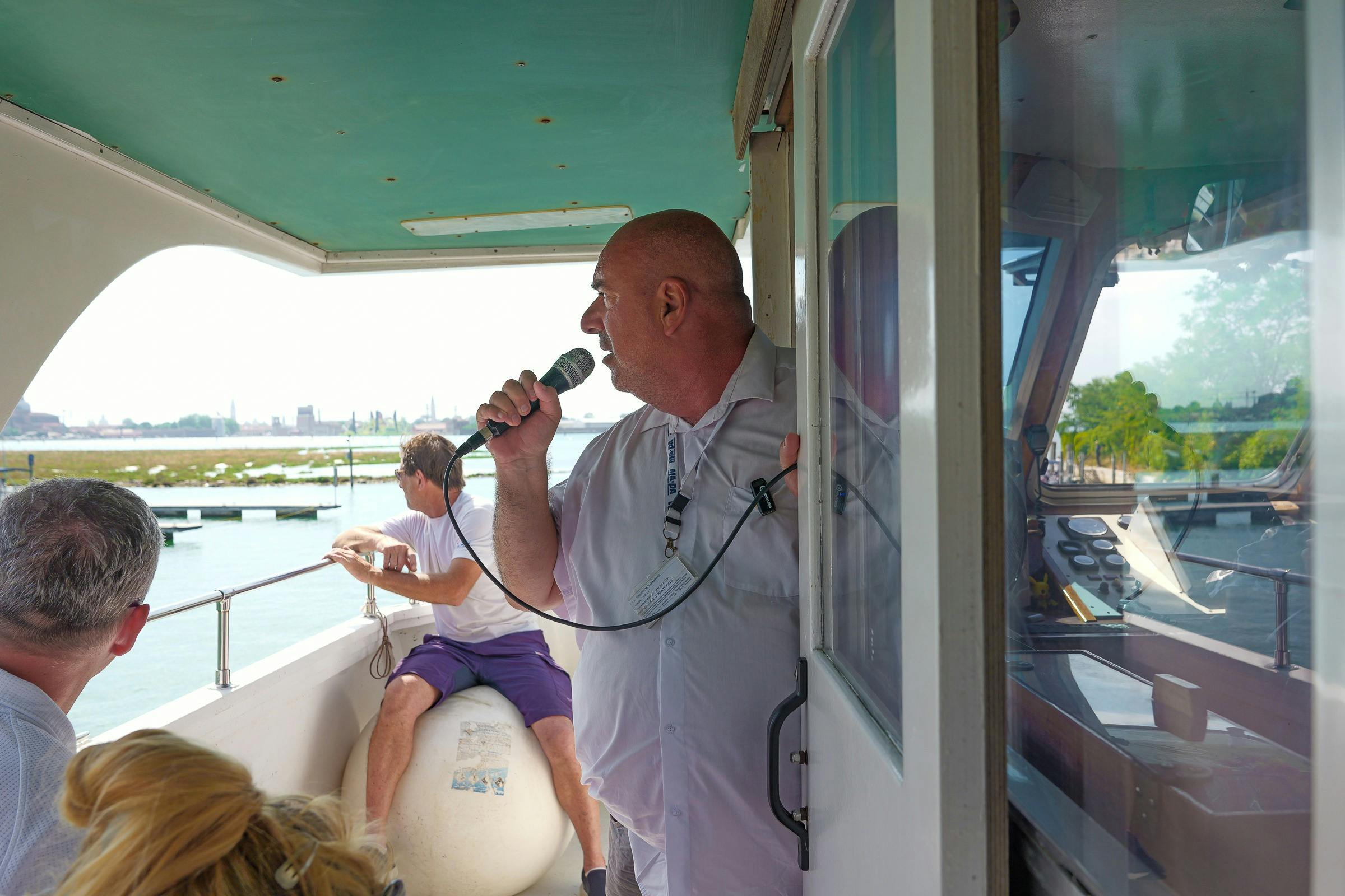 En mand, der holder en mikrofon på en båd med en anden mand siddende bag sig, med vand og fjerne bygninger synlige.