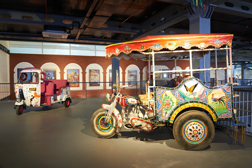 Colorful, decorative auto rickshaw and tuk-tuk on display in an indoor setting with artworks on the wall and an arched window.
