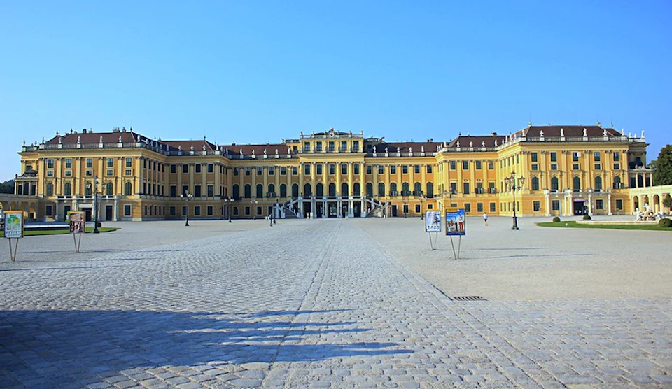 A large, yellow palatial building with numerous windows and an open courtyard with cobblestone paving under a clear blue sky.