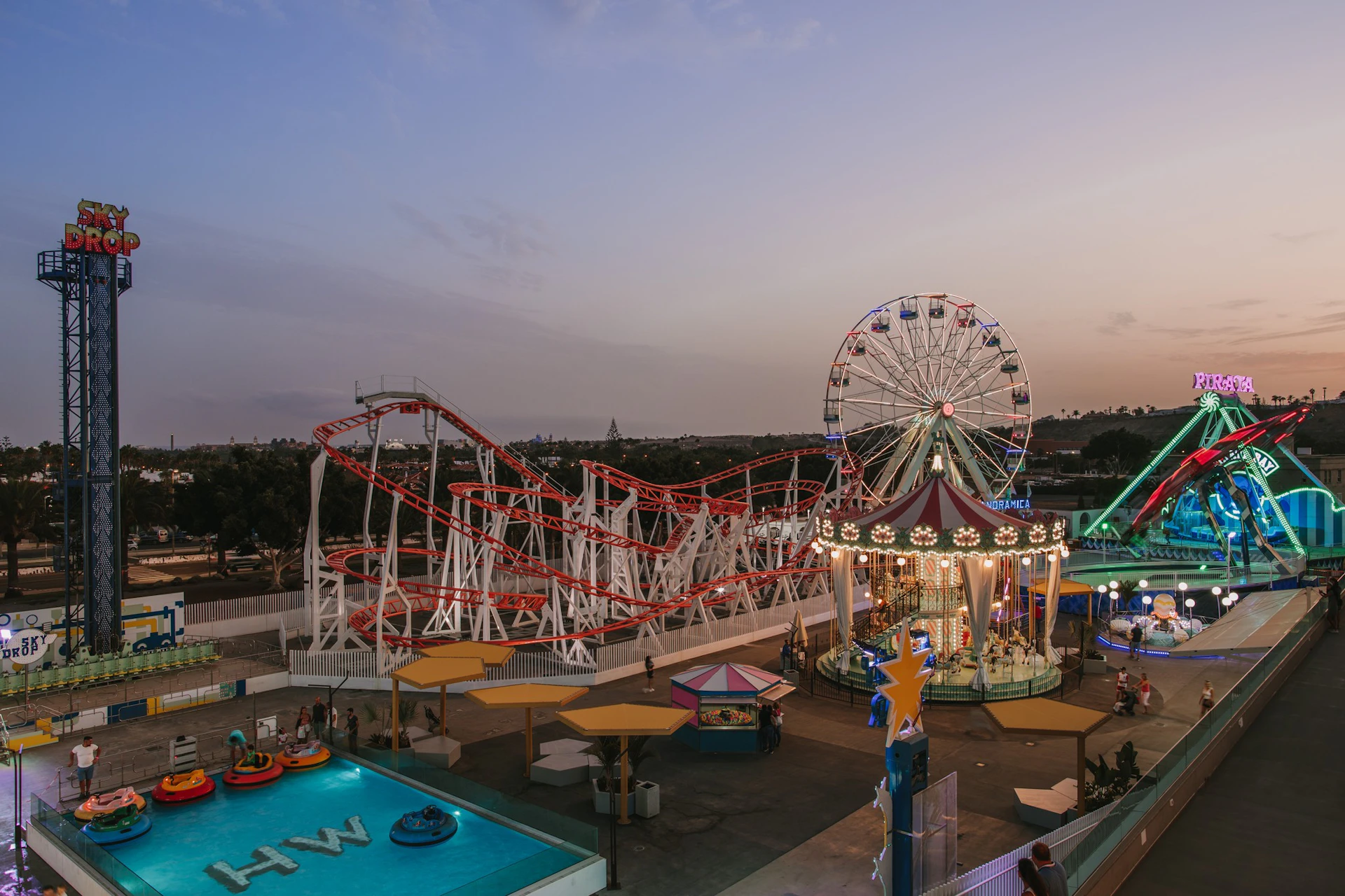 An amusement park at dusk with a rollercoaster, Ferris wheel, carousel, bumper boats, and colorful lights illuminating the area.