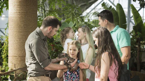 A group of people, including children, interact with a snake held by a man in a lush indoor environment.