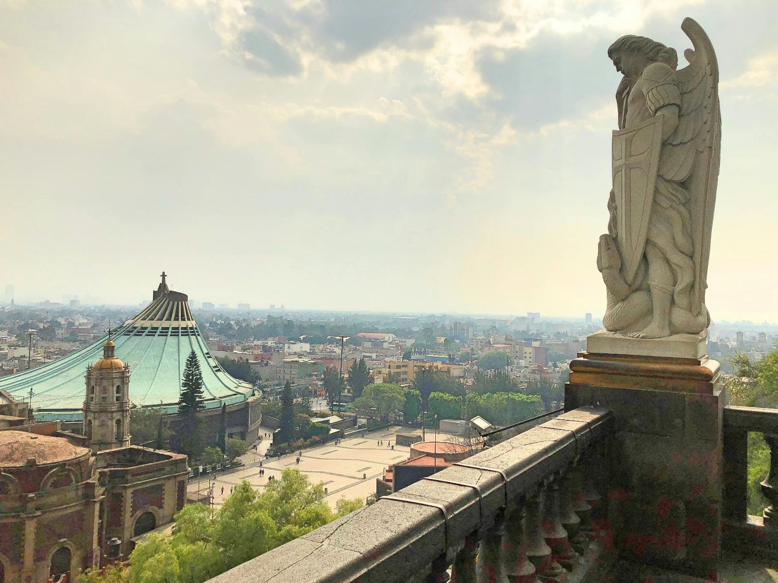 Panoráma města s kupolí se zelenou střechou, okolními budovami a sochou postavy se štítem v popředí.