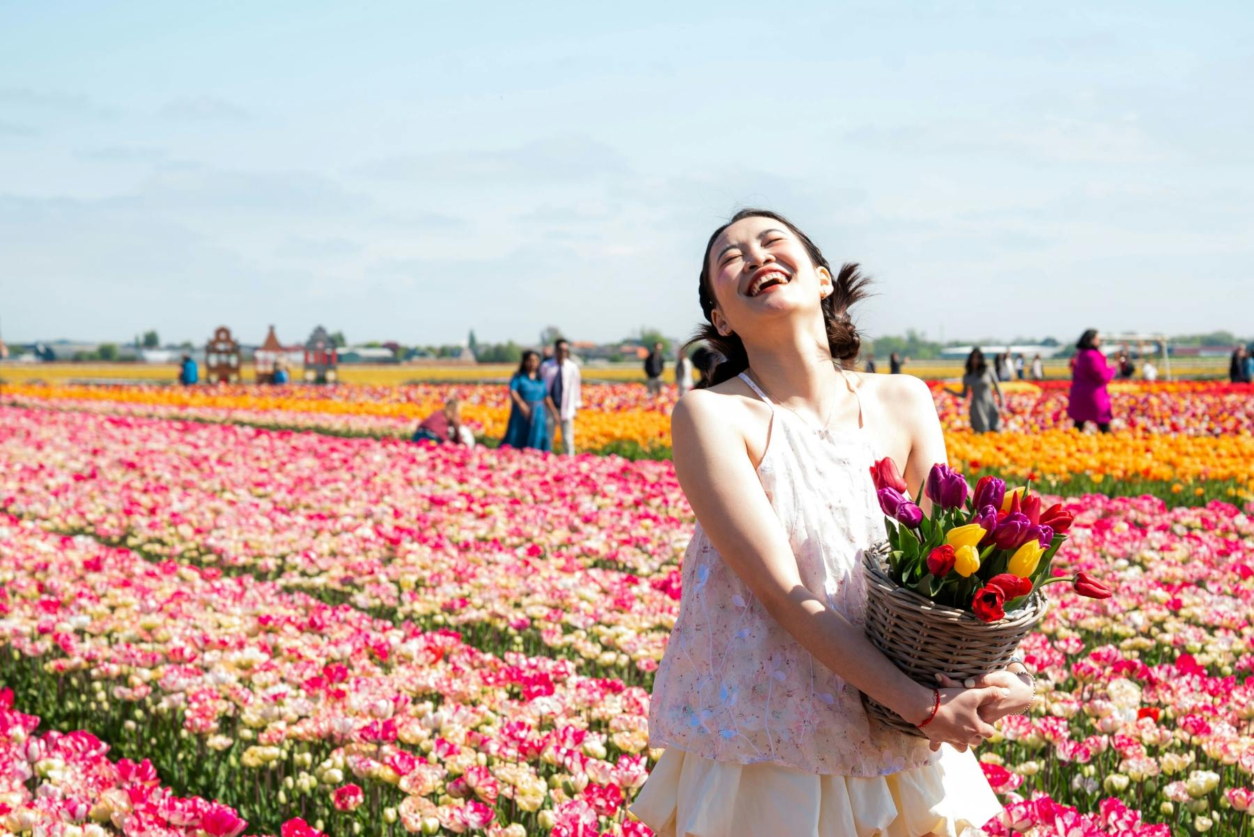 A woman smiling and holding a basket of tulips in a colorful flower field on a sunny day.