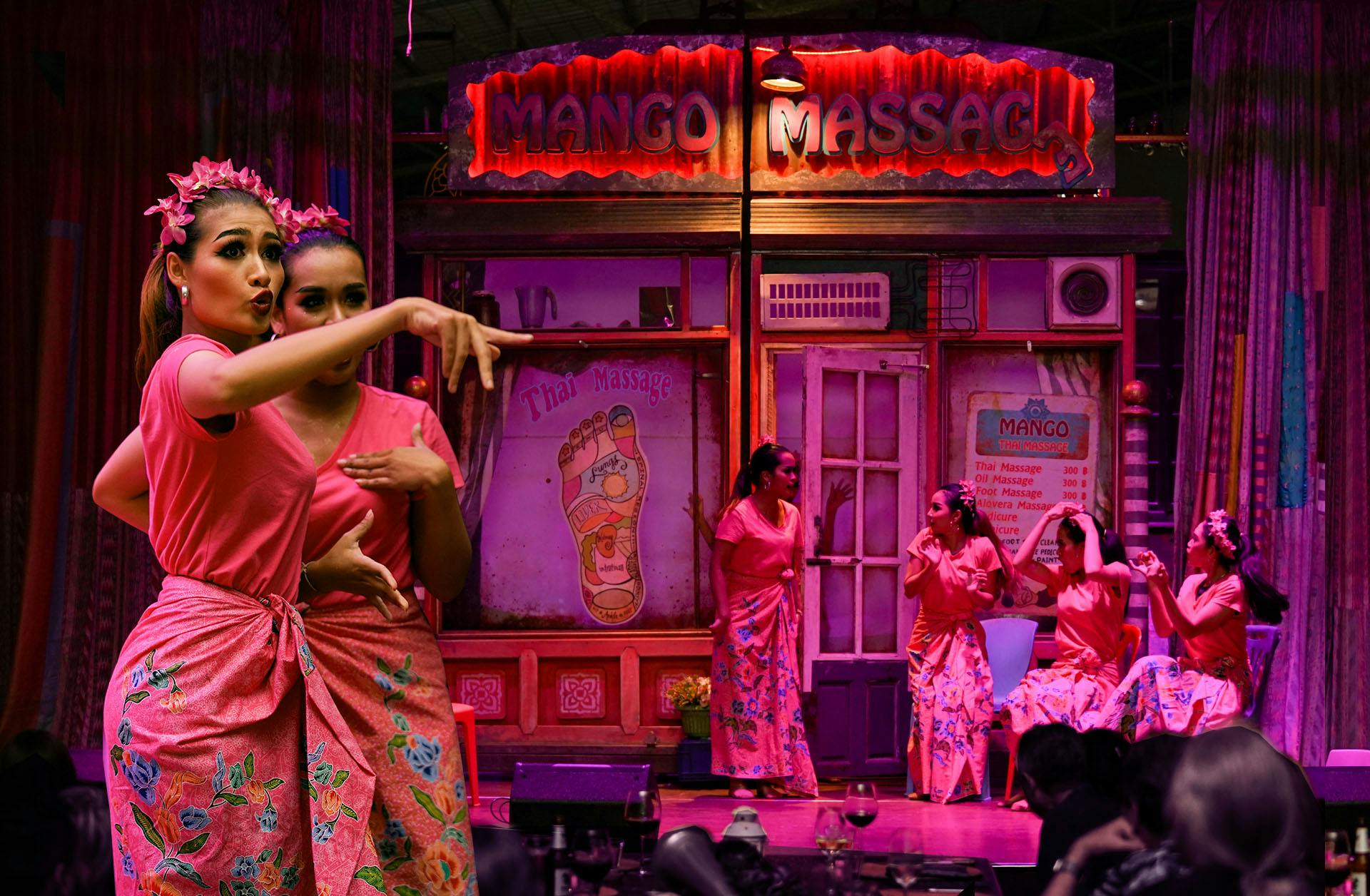 Two performers in pink outfits with floral patterns pose on a stage in front of a brightly lit Thai-themed storefront.