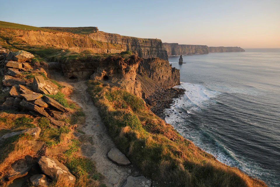 A coastal cliff path at sunset, overlooking the ocean and steep cliffs with a sea stack in the distance.