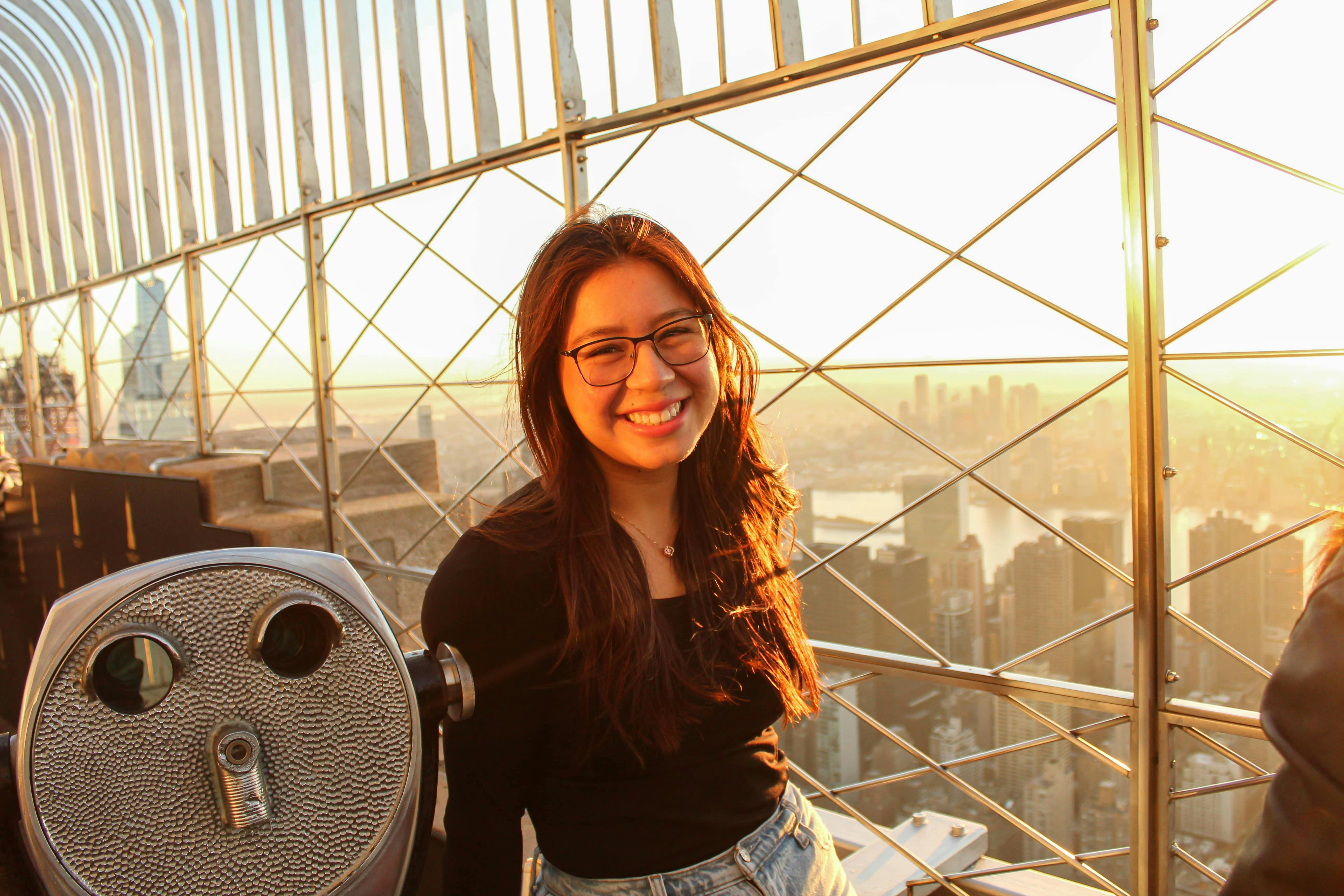 A person smiling on an observation deck with a cityscape and sunset in the background.