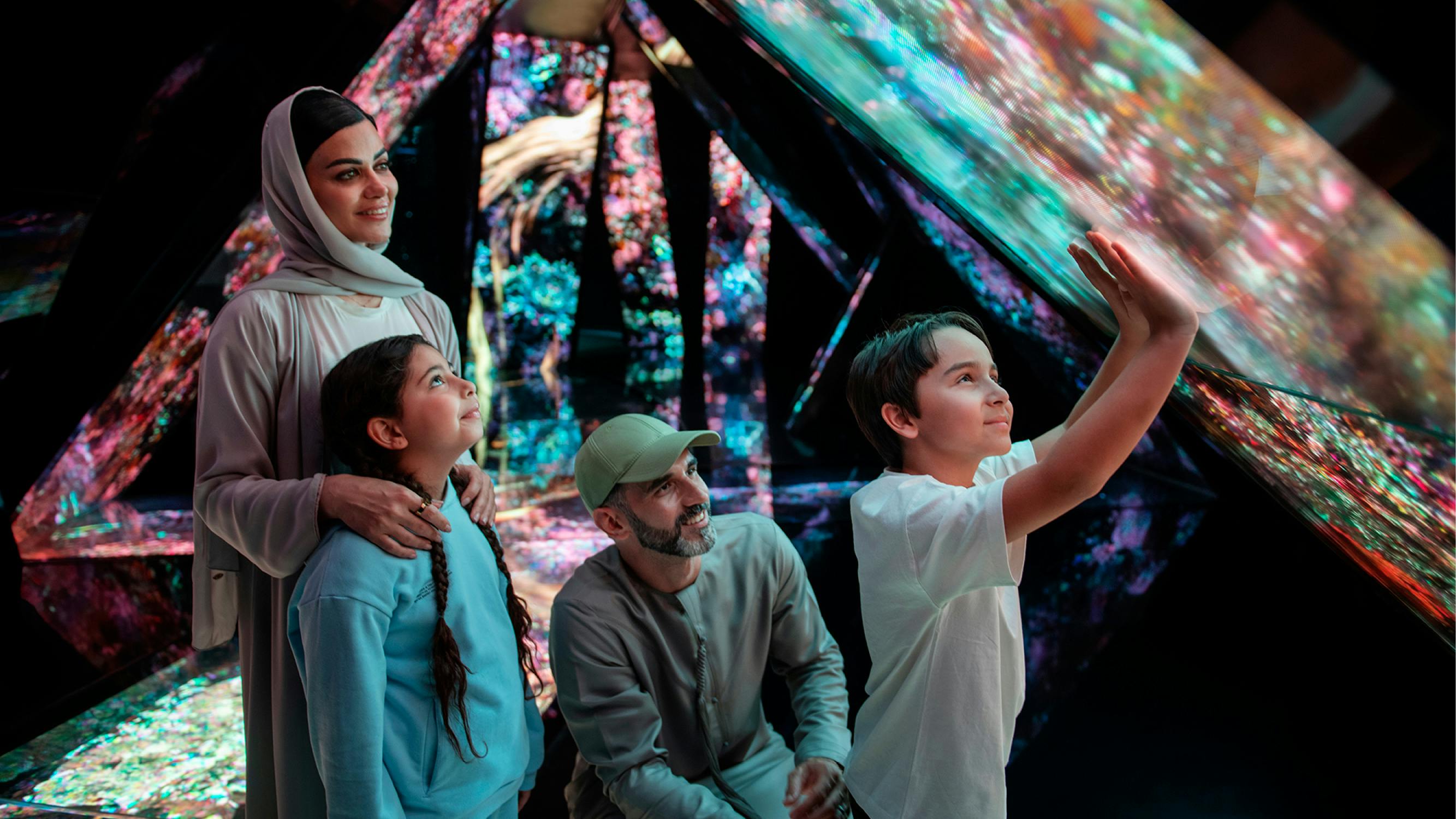 Μια γυναίκα, ένας άνδρας και δύο παιδιά εξερευνούν μια πολύχρωμη, διαδραστική έκθεση με προβαλλόμενη ψηφιακή τέχνη στα γύρω πάνελ.