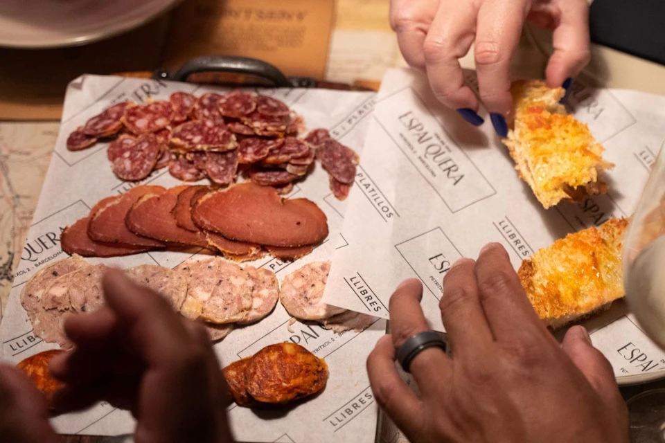 Hands reaching for sliced cured meats and bread on a wooden board lined with branded paper.