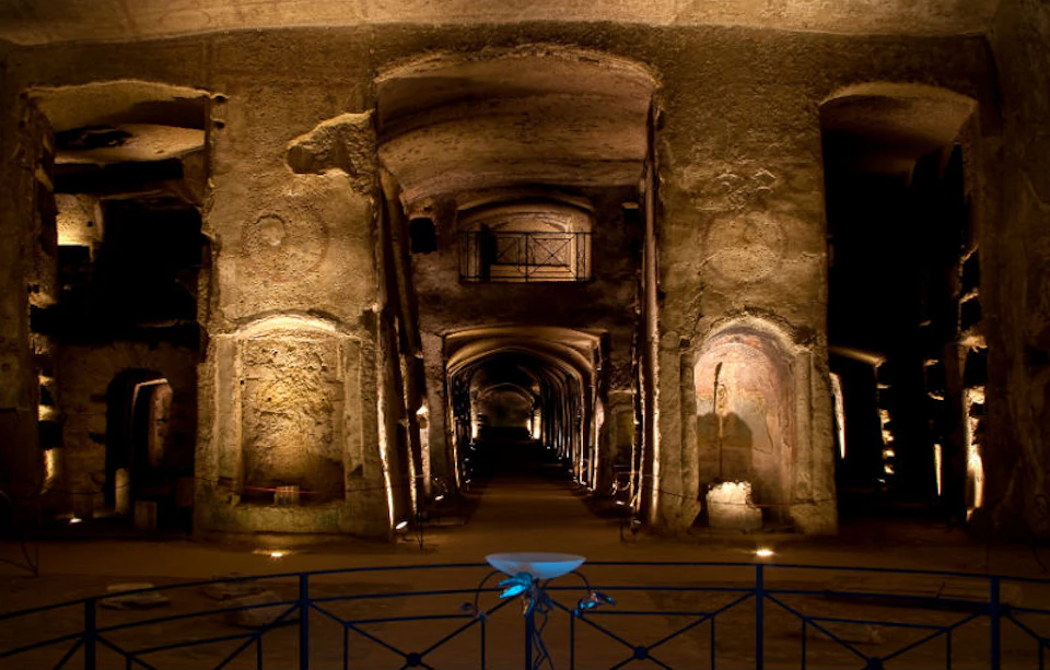 Ancient underground chamber with illuminated archways, columns, and wall carvings, viewed from a blue railing.