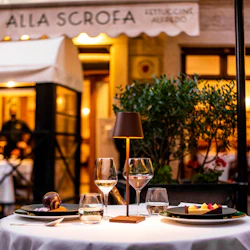 Elegant utomhusrestaurangbord med desserter, vinglas och en liten lampa, framför en entré med en upplyst skylt och krukväxt.