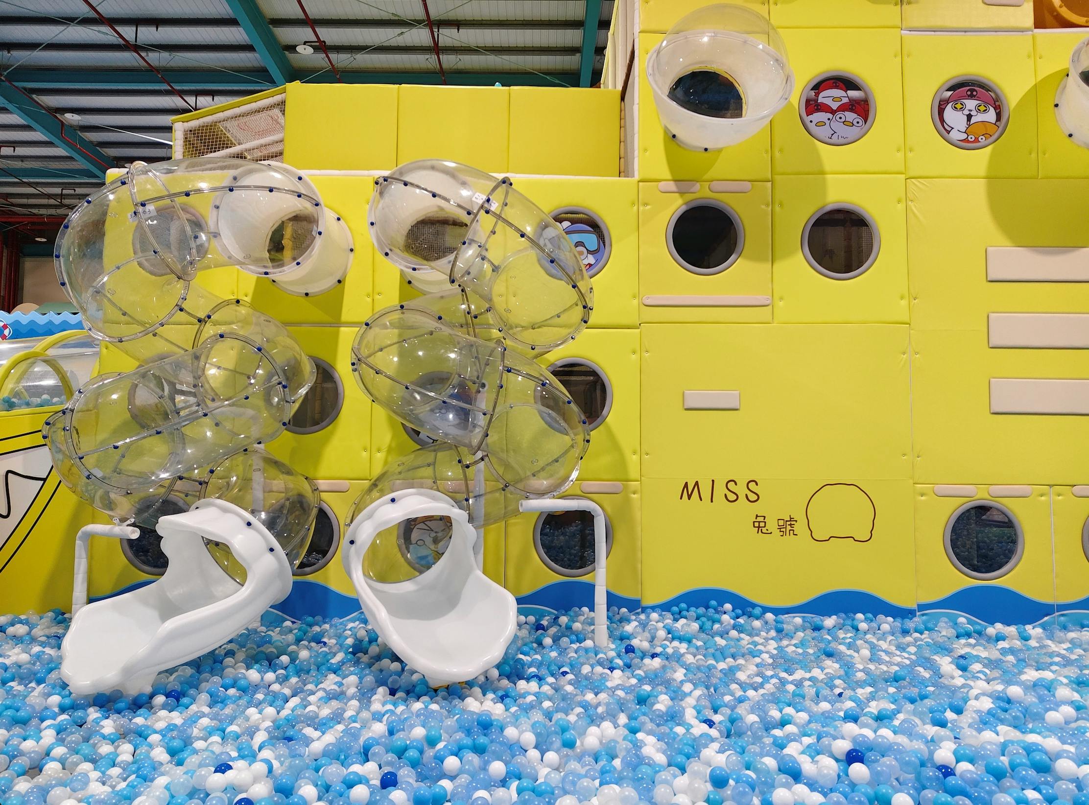 Indoor playground with yellow walls, circular windows, clear plastic climbing structures, white slides, and a ball pit.