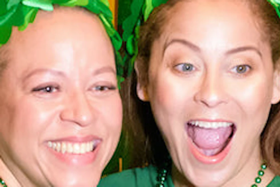 Two women wearing green shirts and shamrock headbands pose excitedly with paper beer props against a Liberty Science Center backdrop.