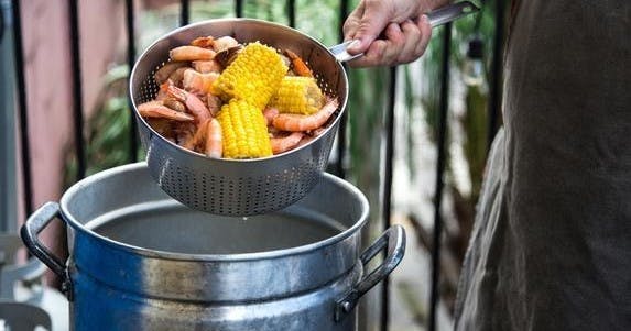 Une main tenant une passoire remplie de crevettes cuites et d'épis de maïs au-dessus d'une grande marmite en métal, à l'extérieur.